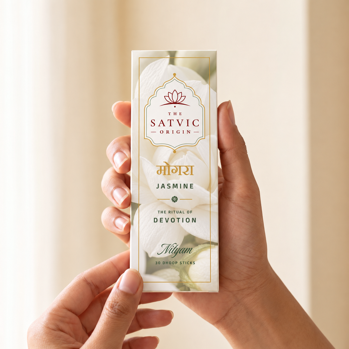 A person's hands holding a cream-colored box of 'The Satvic Origin' Jasmine ritual sticks against a soft, warm beige background.