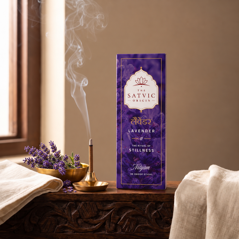 A purple box of The Satvic Origins Lavender incense sticks stands on a carved wooden table, with a lit incense stick burning in a brass holder beside lavender flowers and draped fabric.
