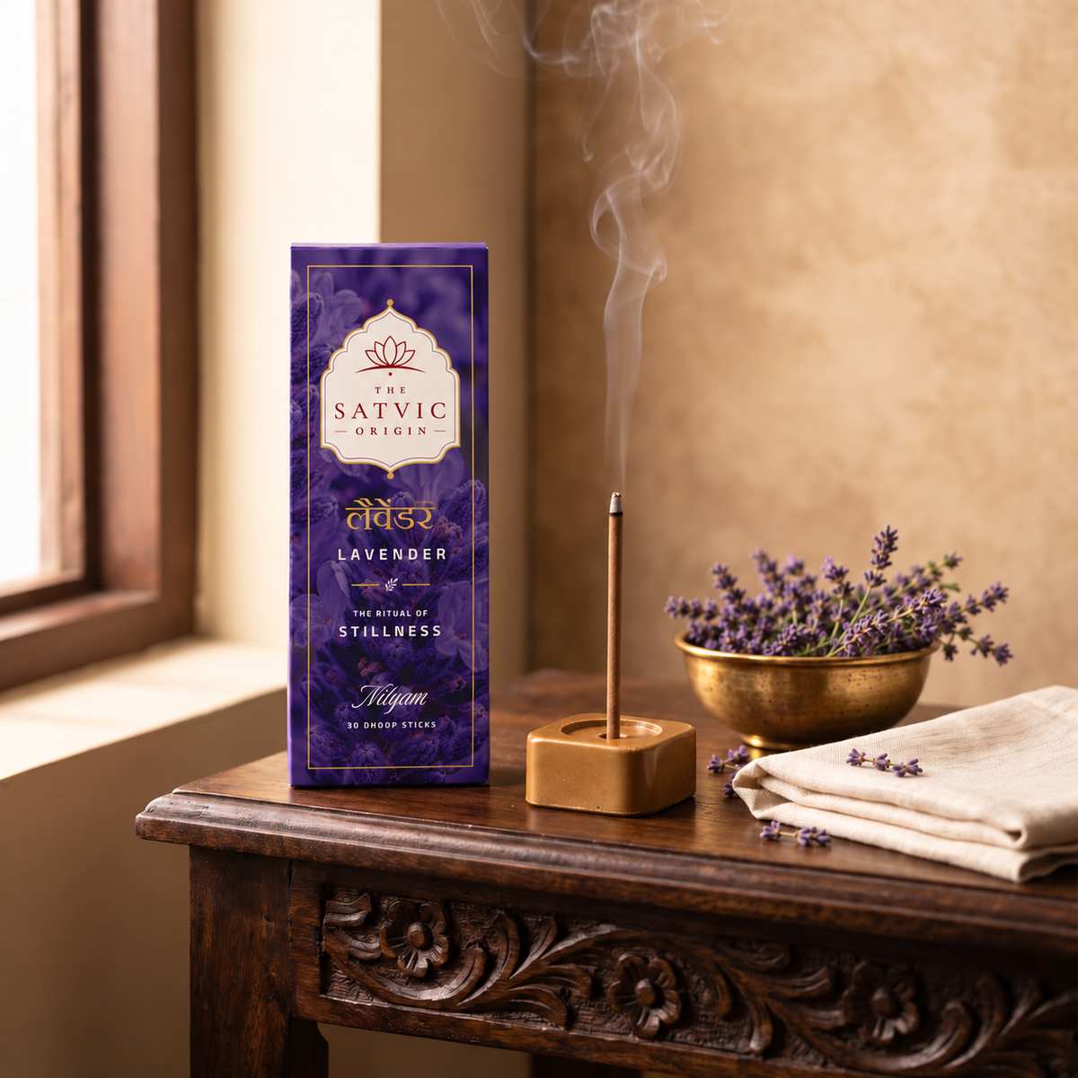 A purple box of The Satvic Origins Lavender Hemp Hash incense sticks sits on a dark wooden table next to a smoking incense stick holder, a gold bowl of lavender, and folded cream cloth in a warm indoor setting.