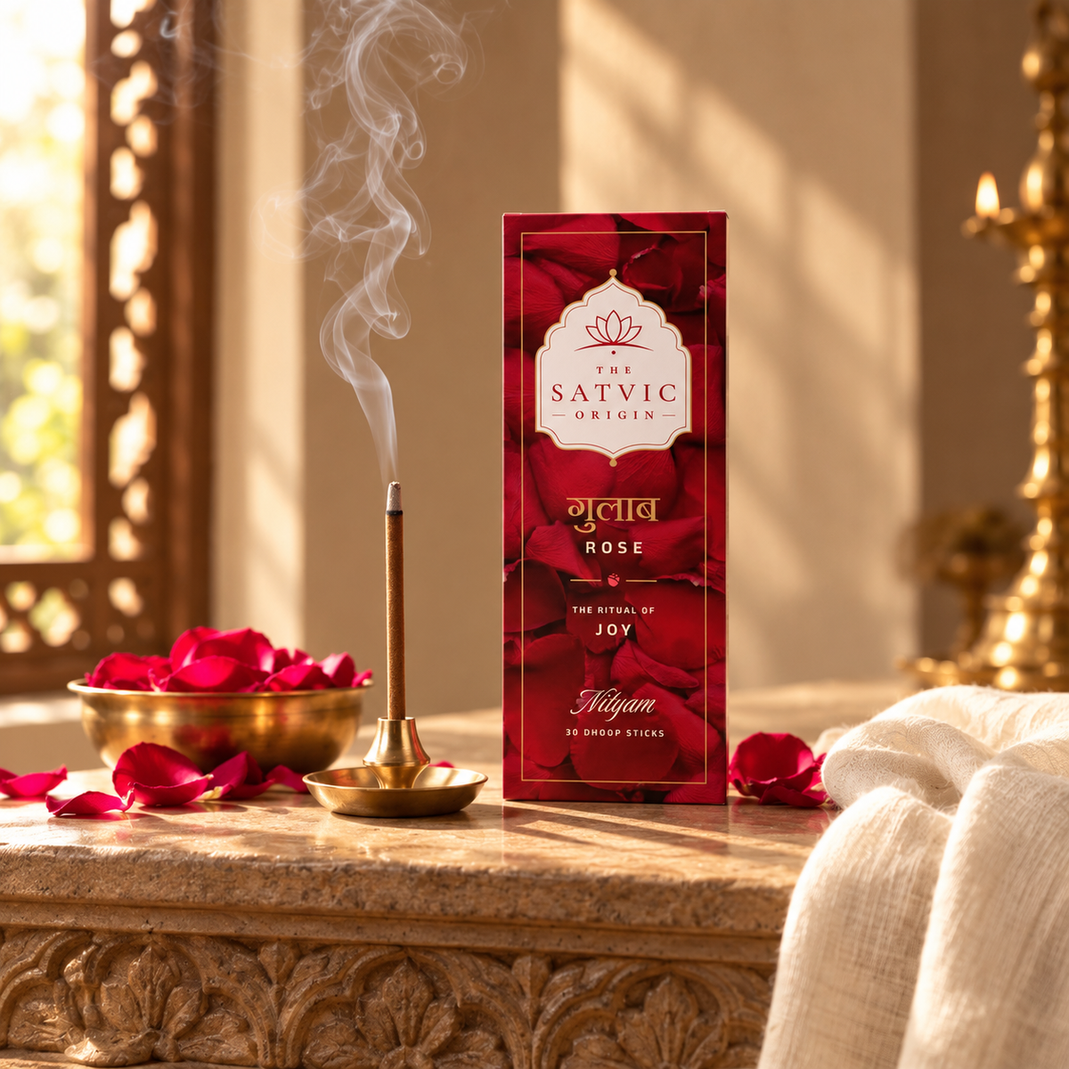 A red rectangular box labeled 'The Satvic Origin' and 'Gulaab Rose' stands on a stone table beside a brass incense burner with smoke rising, scattered red rose petals, and a draped cream cloth in a warm indoor setting.