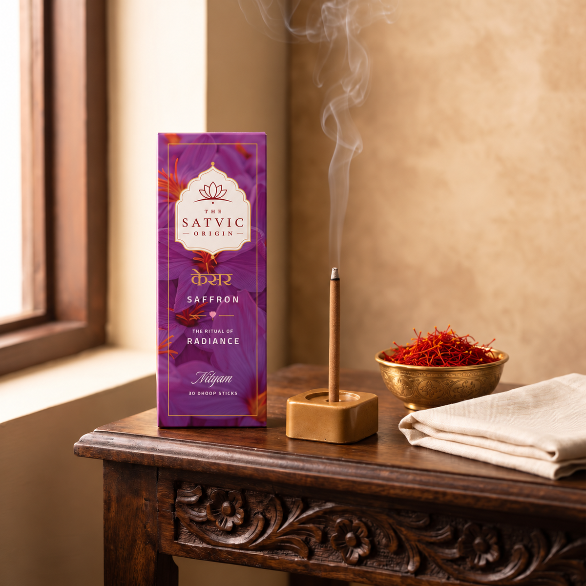 A purple box of Satvic Origin Saffron incense sticks sits on a carved wooden table next to a burning incense stick in a wooden holder, with a small gold bowl of saffron threads and folded white cloth in a warm indoor setting.
