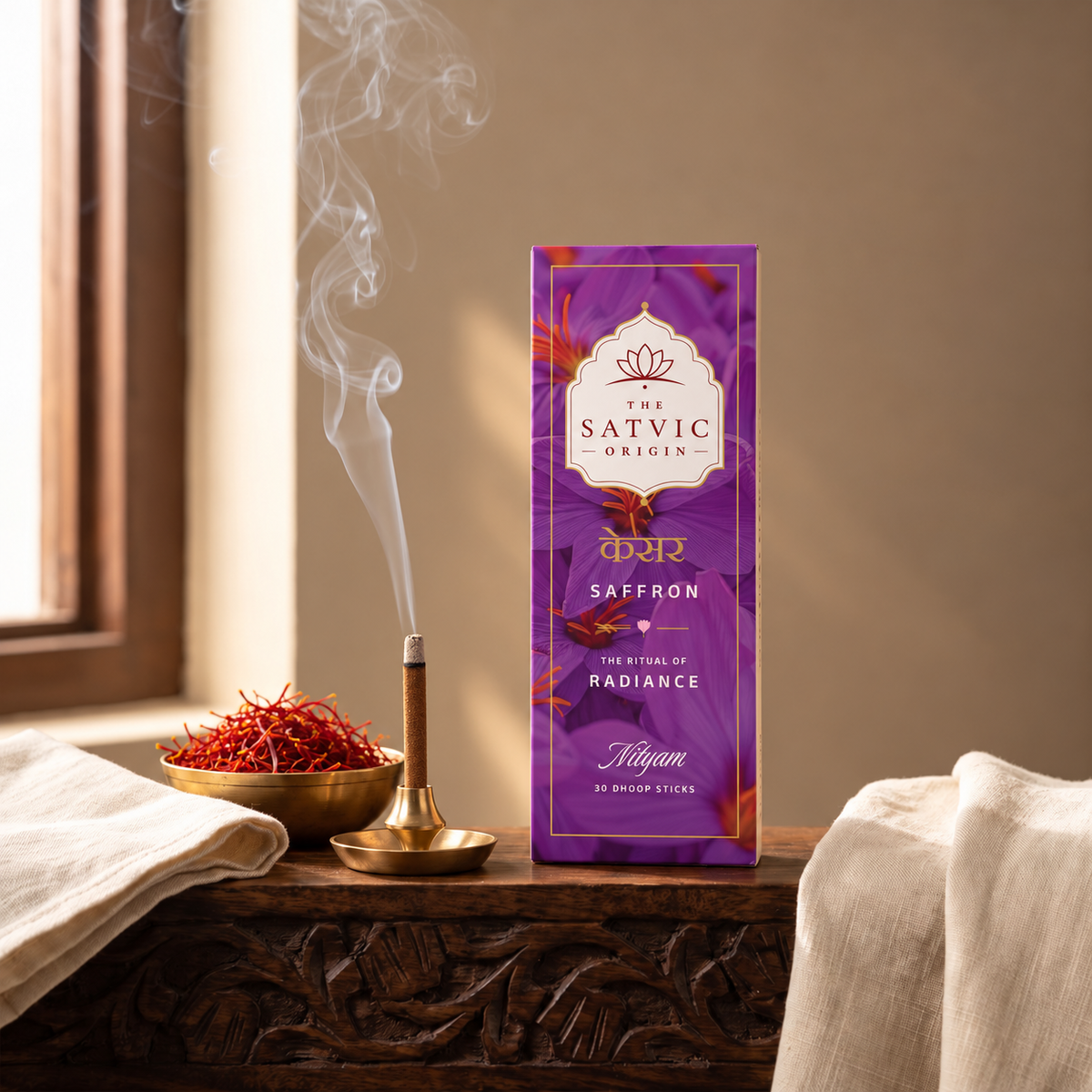 A purple box of Satvic Origin Saffron incense sticks stands on a carved wooden table beside a burning incense holder, a bowl of saffron threads, and draped white fabric, with warm sunlight and window light in the background.