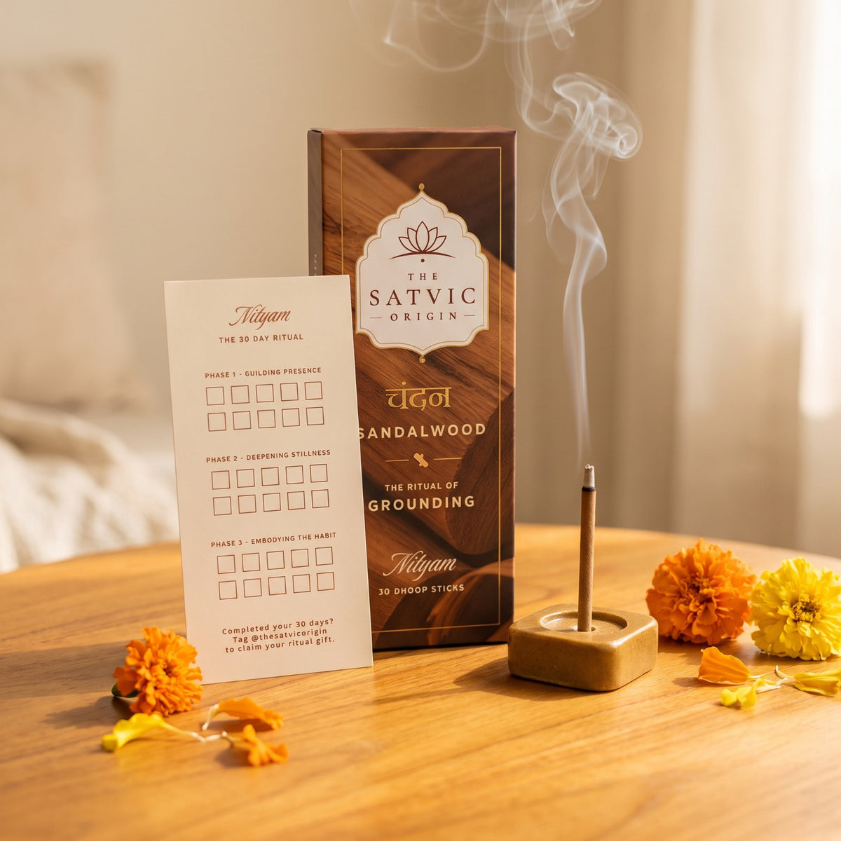 A boxed incense set labeled 'The Satvic Origin' stands on a wooden table beside a lit incense stick in a wooden holder, with white smoke rising. A printed ritual card is also visible.