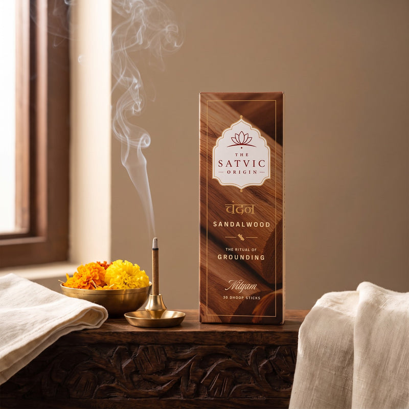 A box of Sandalwood incense from The Satvic Origin stands on a carved wooden table beside a brass incense burner with rising smoke, a small bowl of marigolds, and draped white fabric in a warm indoor setting.