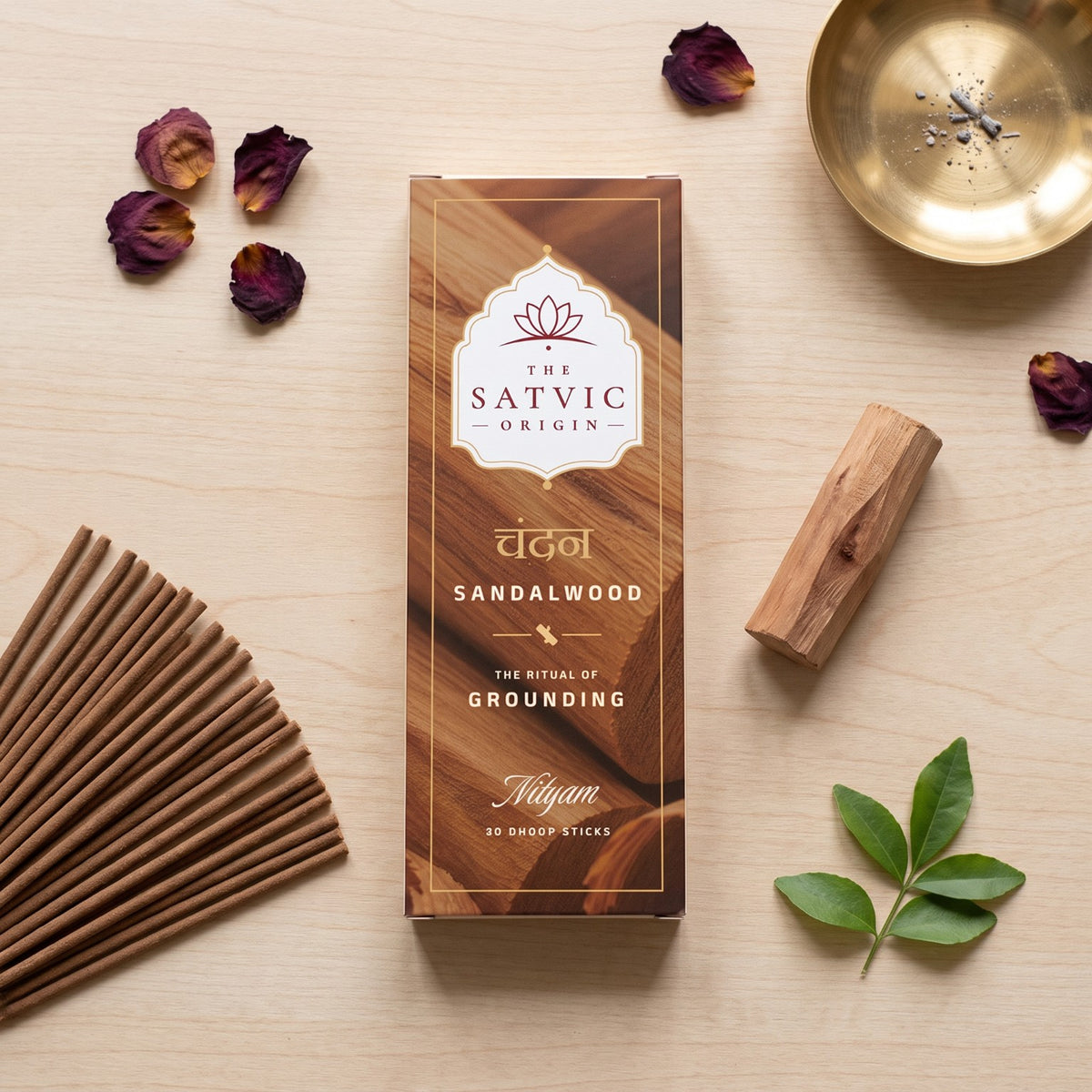 A boxed incense stick set from The Satvic Origin, labeled Sandalwood, arranged in a styled flat lay on a light wood surface with dried rose petals, a brass plate, green leaves, and additional incense sticks.