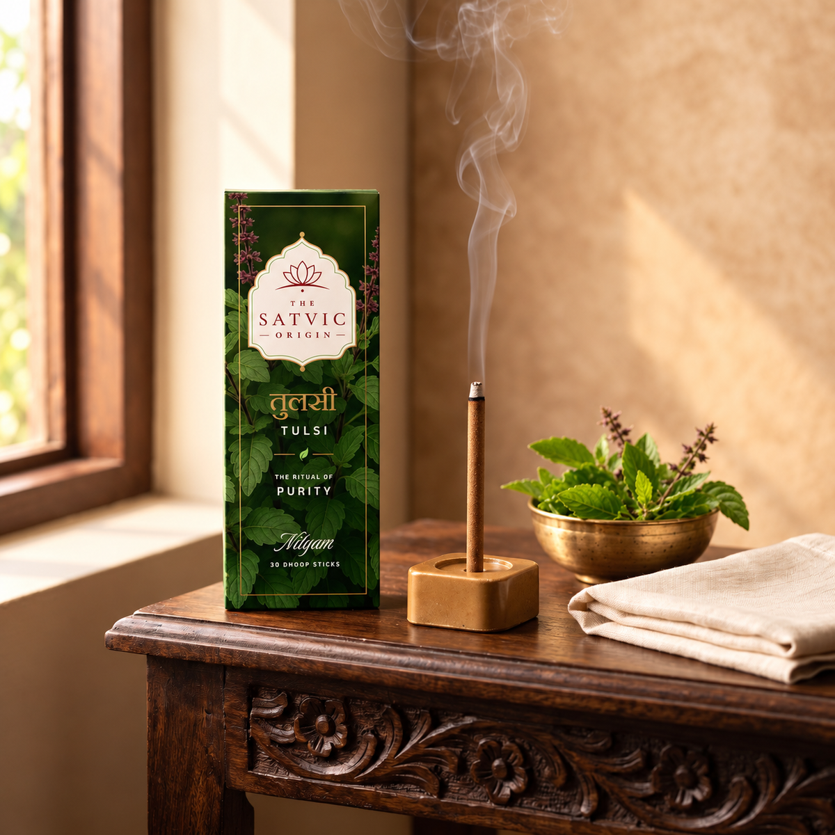 A box of The Satvic Origins Tulsi incense sticks stands on a carved wooden table beside a smoking incense stick holder, a small gold bowl of fresh tulsi leaves, and a folded cream cloth in a warm indoor setting.