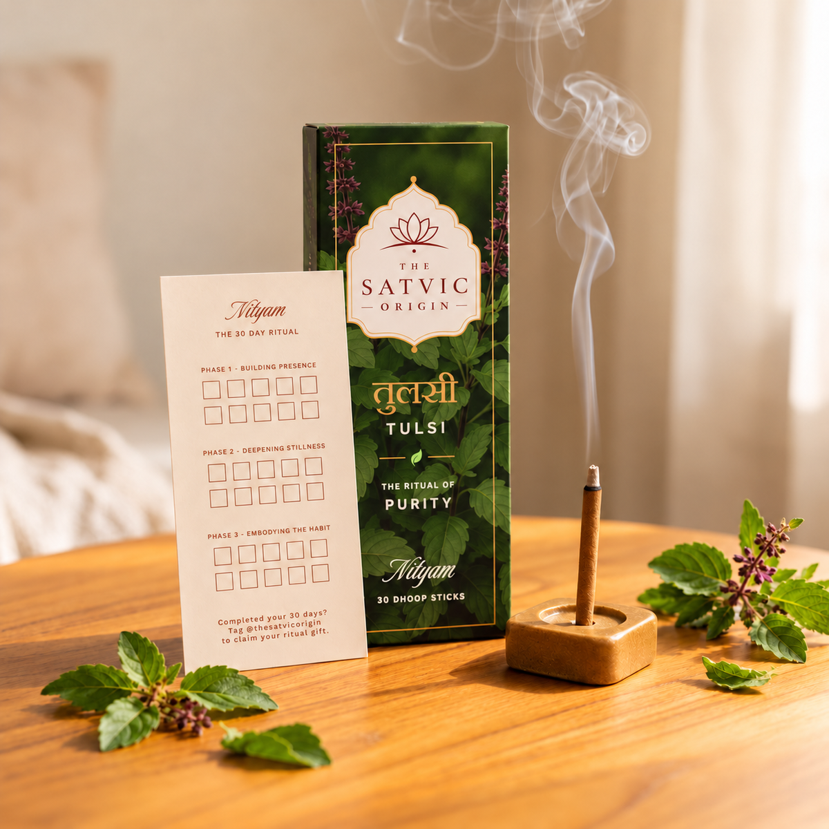 A smoking incense stick in a wooden holder beside a green box labeled 'The Satvic Origin Tulsi' and a cream-colored ritual card, arranged on a wooden table with tulsi leaves and flowers in a softly lit indoor setting.